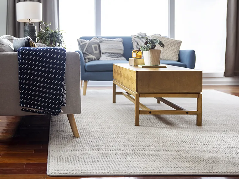 Custom-Bound-Rugs Light textured area rug over hardwood flooring in a living room with a blue sofa and wooden coffee table.